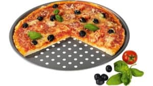 Eine runde Pizza mit Käse, schwarzen Oliven und Basilikum liegt auf einer Gourmetmaxx Grillplatte, ein großes Stück fehlt. Tomaten, Basilikum und Oliven sind ordentlich neben der Platte angeordnet.